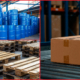 Industrial packaging setup showing blue chemical drums, stacked wooden pallets, and a sealed corrugated box in a warehouse.